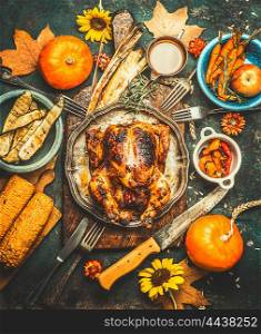 Roasted whole stuffed chicken or turkey for Thanksgiving Day , served with sauce, pumpkins, corn and autumn harvest vegetables, kitchen knife and cutlery on dark rustic background, top view