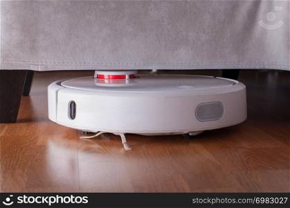 Robotic vacuum cleaner runs under sofa in room on laminate floor. Robot controlled by voice commands to direct cleaning. Modern smart cleaning technology housekeeping.. Robotic vacuum cleaner runs under sofa in room on laminate floor. Robot controlled by voice commands to direct cleaning. Modern smart cleaning technology housekeeping