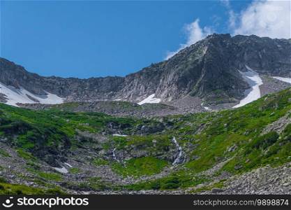 Rock ridge with snow and stone placers under blue sky. Summer trip to mountain valley. Atmospheric alpine landscape with stony meadow with green grass.
