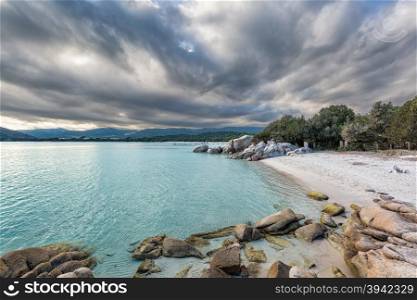 Rocks and boulders in a translucent turquoise sea with a jetty in the background and cloudy skies at Santa Giulia beach in Corsica