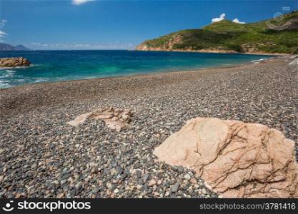 Rocks and pebble beach at Plage de Bussaglia against a turquoise sea with maquis covered rocks and clouds in background