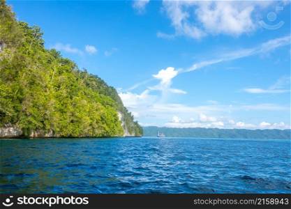 Rocky coast of the tropical island of Indonesia in sunny weather. Rainforest on the slope. Yacht at a distance. Sunny Rocky Shores with Rainforest and Yacht in the Distance