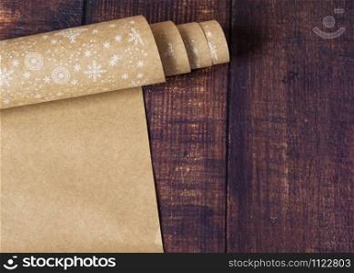 Roll of brown kraft paper with a pattern of snowflakes on a wooden background. Eco paper. Secondary, environmentally friendly. Material for package packaging. Kraft paper for gift wrapping.. Roll of brown kraft paper with a pattern of snowflakes on a wooden background. Eco paper. Secondary, environmentally friendly. Material for package packaging. Gift wrapping paper.