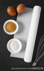 Roll of white baking paper with whisk and baking ingredients such as flour, sugar and eggs on the side, photographed overhead on slate with natural light