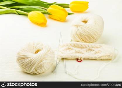 roll of white soft knitting yarn, knitting mittens and yellow tulips on lighten background