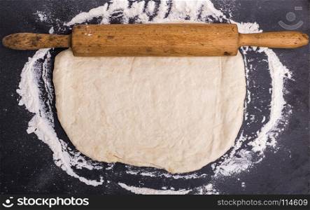 rolled dough of white wheat flour with a wooden rolling pin on a black surface