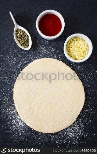 Rolled out pizza dough on floured slate surface with ingredients, such as tomato sauce, oregano and grated cheese on the side, photographed overhead with natural light