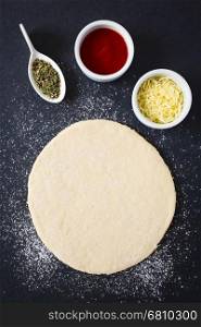 Rolled out pizza dough on floured slate surface with ingredients, such as tomato sauce, oregano and grated cheese on the side, photographed overhead with natural light