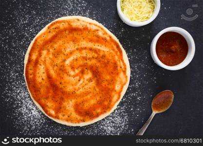 Rolled out pizza dough with tomato sauce on floured slate surface, tomato sauce and grated cheese on the side, photographed overhead with natural light
