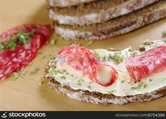 Rolled salami slice filled with cream cheese with dried herbs on a slice of brown bread on wooden board and ingredients in the background (Selective Focus, Focus on the front of the salami roll and filling). Rolled Salami on Brown Bread