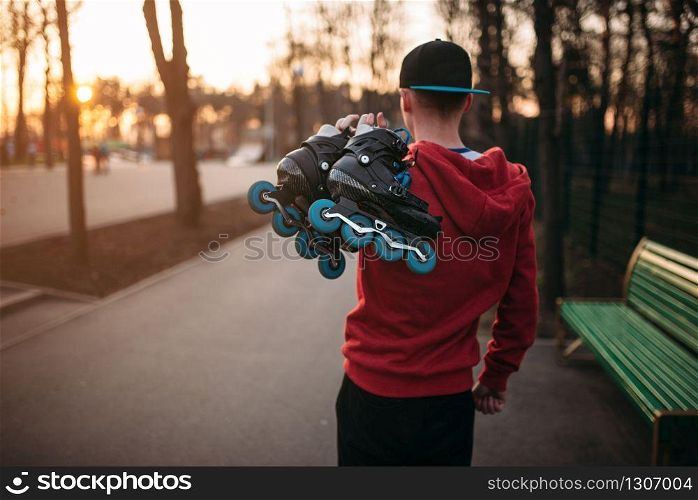 Roller skater with skates in hands on sidewalk in city park, back view. Male rollerskater leisure