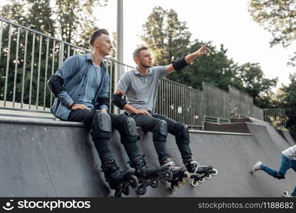 Roller skating, two male skaters sitting on the ramp in park. Urban roller-skating, active extreme sport outdoors. Roller skating, two skaters sitting on the ramp