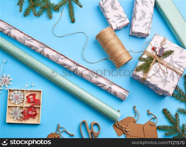 rolls with wrapping paper, brown rope, scissors, decor and a wrapped square box with a gift. DIY gift making process, blue background