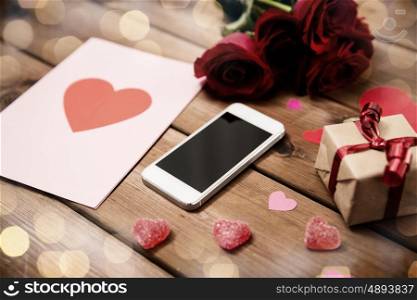 romance, valentines day and holidays concept - close up of smartphone, gift box, red roses and greeting card with heart-shaped candies on wood (vintage effect)
