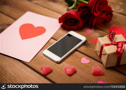 romance, valentines day and holidays concept - close up of smartphone, gift box, red roses and greeting card with heart-shaped candies on wood