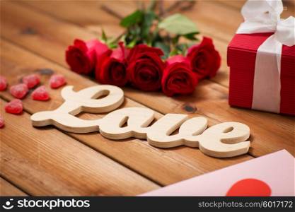 romance, valentines day and holidays concept - close up of word love, gift box, red roses and greeting card with heart-shaped candies on wood