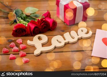 romance, valentines day and holidays concept - close up of word love, gift box, red roses and greeting card with heart-shaped candies and golden lights on wood