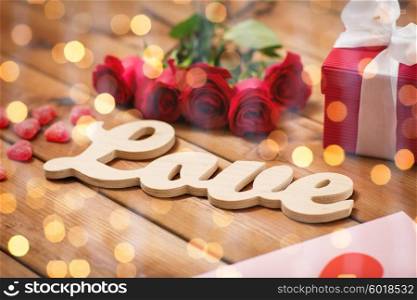 romance, valentines day and holidays concept - close up of word love, gift box, red roses and greeting card with heart-shaped candies on wood over lights background