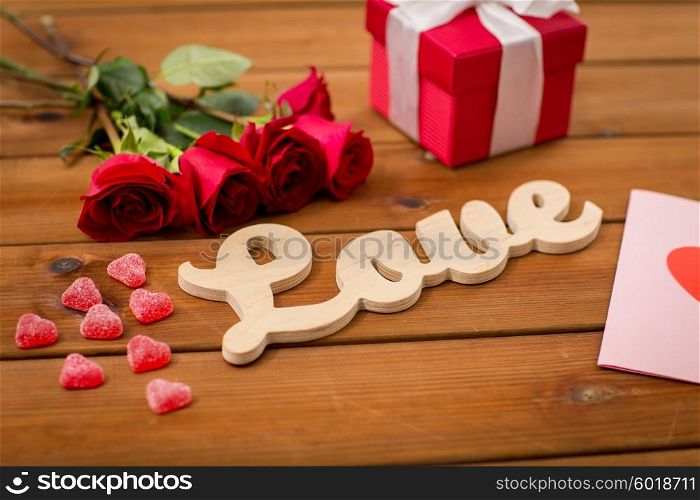 romance, valentines day and holidays concept - close up of word love, gift box, red roses and greeting card with heart-shaped candies on wood