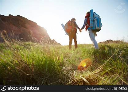 Romantic adventure of young tourist couple, low angle of man and woman going uphill holding hands towards bright sunlight on hiking path in mountains