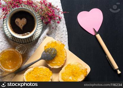 Romantic breakfast with flowers, heart shaped sticky note, bread with jam on a trencher and a cup of coffee with a foam heart, on a black table.