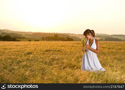 Romantic brunette woman in sunset corn field wear white dress, holding bouquet of flowers