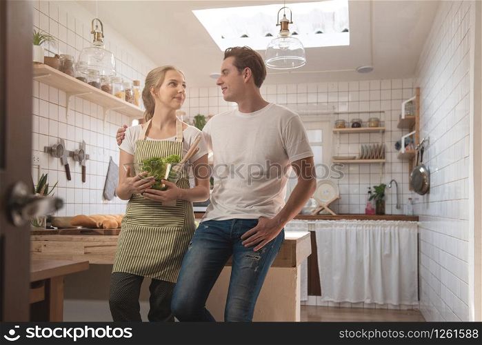 Romantic caucasian couple in love having great time together in the kitchen.Happy young man and woman in kitchen hand hold blow of salad and look at each other with smiling face.