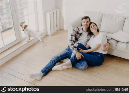Romantic couple enjoy togetherness, embrace and feel support of each other, sit on floor near sofa, glad to spend free time together, dressed casually, have own pet. Husband and wife in living room