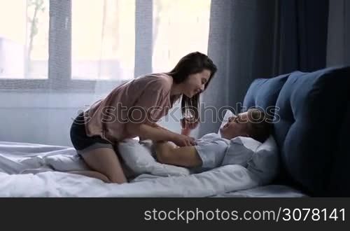 Romantic couple in love lying on bed and enjoying time together after waking up. Beautiful brunette woman waking her boyfriend, tickling, playing and kissing him while man lying on bed in comfortable bedroom.