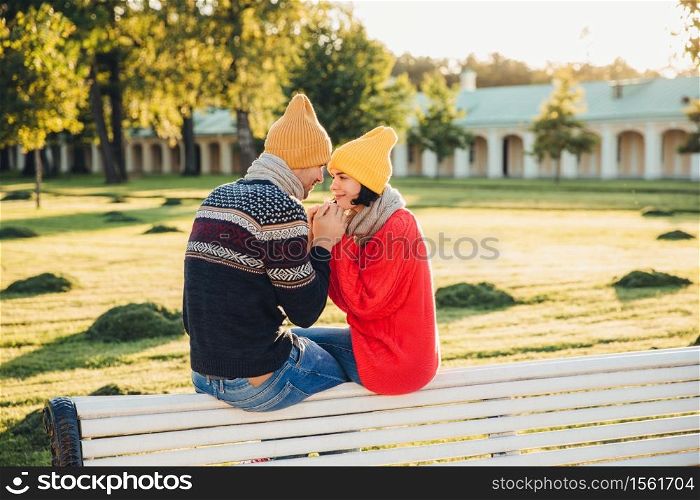 Romantic couple sit on bench, enjoy sunny day, keep hands together, look with great love at each other, have good relationships. Female has date with boyfriend in park, admire beautiful nature