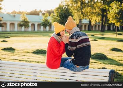 Romantic couple sit on bench, enjoy sunny day, keep hands together, look with great love at each other, have good relationships. Female has date with boyfriend in park, admire beautiful nature