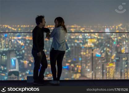 Romantic Couple standing on the rooftop from bangkok modern building at night time over the photo blurred of cityscape background, Lover and calentine holiday concept, low light