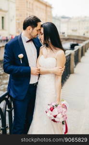 Romantic couple stands on bridge stand close to each other, look affectionately at each other, feel love and passion. Handsome bridegroom and bride in wedding clothes express their feelings.