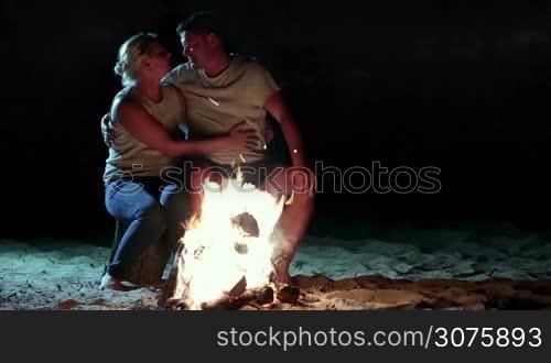 Romantic evening. Charming couple sitting near a campfire at night in the camp on the river bank during summer vacation