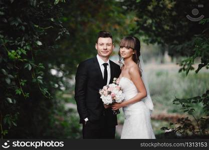 Romantic happy couple newlyweds, bride and groom stands and holding bouquet of pink and purple flowers and greens, greenery with ribbon in the garden. Wedding ceremony on nature.