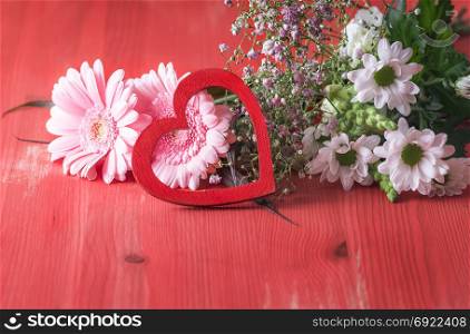 Romantic image with a bouquet of different chrysanthemum flowers and a decorative wooden heart leaned against them, displayed on a red wooden background.