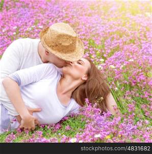 Romantic kisses outdoors, attractive man kissing his beautiful young wife lying down on pink floral meadow, spring time concept