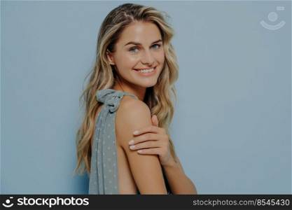 Romantic portrait of young beautiful girl in profile with wavy blond hair in soft blue elegant dress in studio background with copy space, smiling tenderly while looking at camera over her shoulder. Young beautiful female in elegant dress smiling tenderly while looking at camera over her shoulder