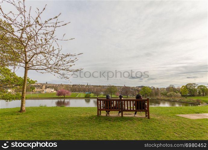 Romantic view of three people sitting on a bench looking towards the pond at Inverleith Park in Edinburgh, Scotland, on a partially cloudy spring afternoon