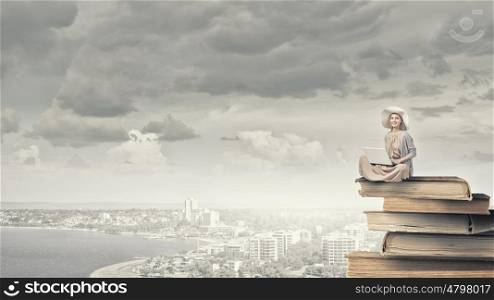 Romantic woman using laptop. Woman in dress and hat sitting on books and working on laptop