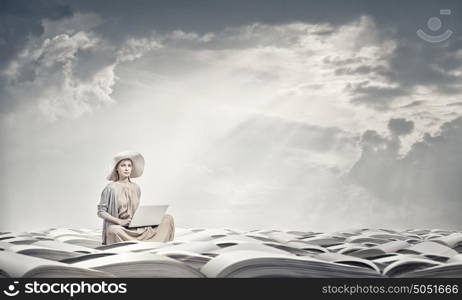 Romantic woman using laptop. Woman in dress and hat sitting on books and working on laptop