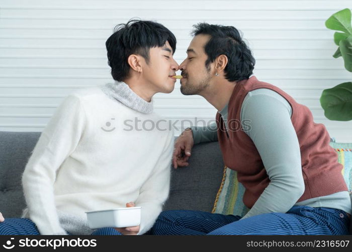 Romantic young Asian Lgbt gay couple eating feeding potato chips mouth to mouth together sitting on sofa at home. Lgbt family lifestyle in winter