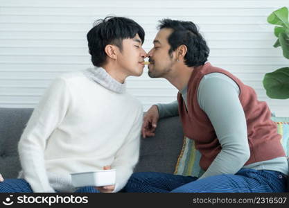 Romantic young Asian Lgbt gay couple eating feeding potato chips mouth to mouth together sitting on sofa at home. Lgbt family lifestyle in winter