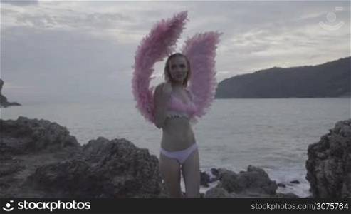 Romantic young beauty as an angel between the rocks. Blonde pretty female woman wearing pink wings, feather bra and bikni bottom smiling while standing on the rocks over sea and sky background - video in slow motion