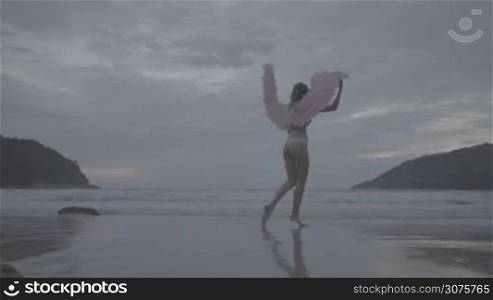 Romantic young beauty as an angel on the beach. Blonde pretty female woman wearing pink wings, feather bra and bikni bottom smiling and walking in the sea over sky background - video in slow motion