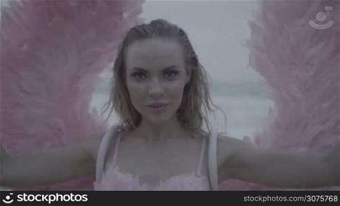 Romantic young beauty as an angel on the beach. Closeup of blonde pretty female woman wearing pink wings and feather bra smiling while standing on the beach over sea and sky background - video in slow motion