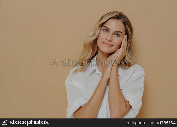 Romantic young blonde lady in casual white shirt keeping folded hands near her face and looking at camera with gentle smile, posing over beige background with copy space. Human face expressions. Romantic young blonde lady in casual white shirt keeping folded hands near her face