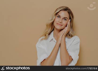 Romantic young blonde lady in casual white shirt keeping folded hands near her face and looking at camera with gentle smile, posing over beige background with copy space. Human face expressions. Romantic young blonde lady in casual white shirt keeping folded hands near her face