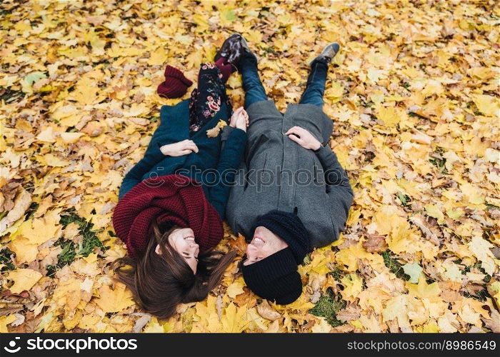 Romantic young couple lie on yellow leaves in park, hold hands together, look at each other with great love, enjoy autumn sunny day. People, relationships, togetherness and romance concept