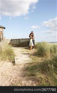 Romantic Young Couple Standing By Wooden Fence Of Beach Hut Amongst Dunes